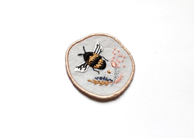 May include: A round embroidered patch with a white background and a brown outline. The patch features a black and yellow bee with white wings flying over a pink flower and green leaves.