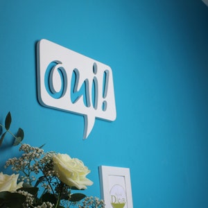 May include: White wooden speech bubble with the word "oui!" in a stylized font, mounted on a bright blue wall. A bouquet of white roses and baby's breath is in the foreground.