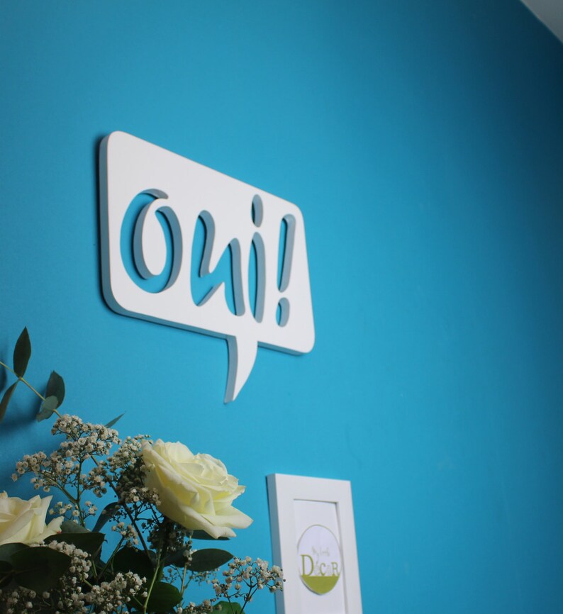 May include: White wooden speech bubble with the word "oui!" in a stylized font, mounted on a bright blue wall. A bouquet of white roses and baby's breath is in the foreground.