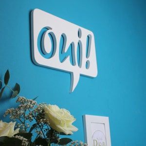 May include: White wooden speech bubble with the word "oui!" in a stylized font. The speech bubble is mounted on a blue wall.