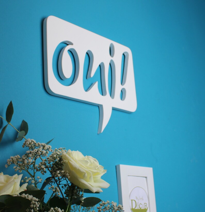 May include: White wooden speech bubble with the word "oui!" in a stylized font. The speech bubble is mounted on a blue wall.