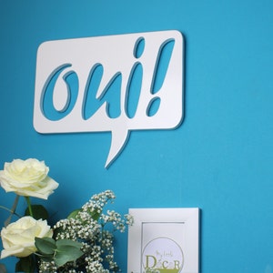 May include: White wooden speech bubble with the word "oui" cut out in a whimsical font. The speech bubble is mounted on a bright blue wall. A bouquet of white roses and baby's breath sits in front of the speech bubble.