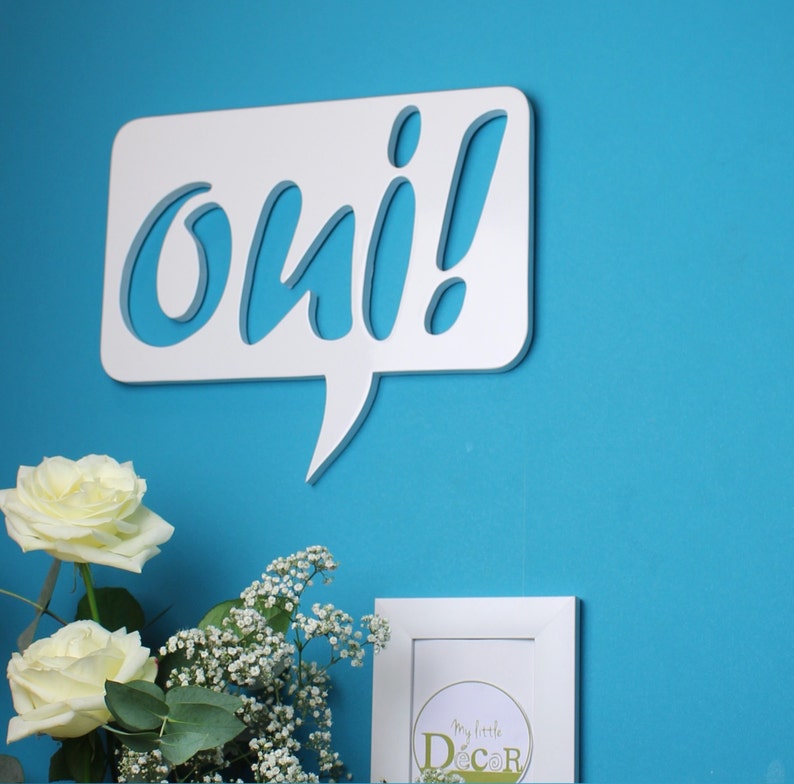May include: White wooden speech bubble with the word "oui" cut out in a whimsical font. The speech bubble is mounted on a bright blue wall. A bouquet of white roses and baby's breath sits in front of the speech bubble.