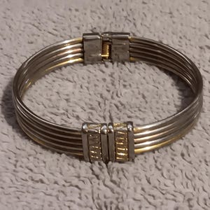 May include: A multi-strand hinged bangle bracelet featuring alternating silver and gold-toned metal wires. The bracelet includes two rectangular metal clasps with a textured, grooved surface that secure the multiple strands together.