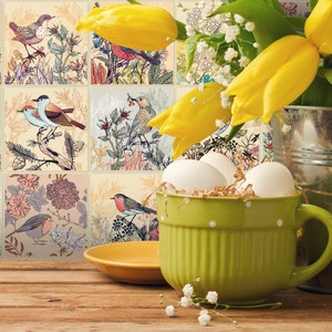 May include: A green ceramic mug with white polka dots filled with white eggs and straw, sitting on a wooden table. Yellow tulips and a silver bucket are in the background. The mug is decorated with a floral pattern and birds.