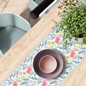 May include: A table setting featuring a floral patterned table runner with pink and blue flowers, a purple plate, and a smaller pink bowl. A potted plant with green leaves sits on the table, next to a light blue chair.