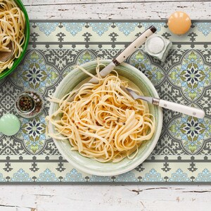 May include: Overhead shot of a table setting with pasta in green and light green bowls. A decorative patterned placemat with blue, green, and gray floral designs is visible. A knife, fork, salt, pepper, and a small orange object are also on the table.