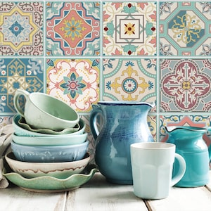 May include: A collection of ceramic tableware, including bowls, cups, and pitchers, in shades of blue and green, arranged on a white wooden surface. The background features a colorful tile pattern with floral and geometric designs.