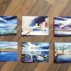 May include: Six small landscape paintings featuring coastal and cityscape scenes. The paintings depict dramatic skies, water, and buildings, with a variety of colors including blues, oranges, and grays. The artworks are displayed on a wooden surface.