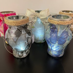 May include: Four glass candle holders with a rope handle and a decorative design. Each holder features a different butterfly design with a light inside. The designs include a blue butterfly, a white butterfly, a black and white butterfly, and a floral design.