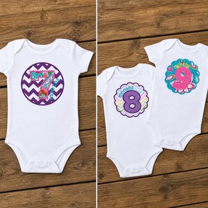 May include: White baby onesies with colorful appliqued numbers 7, 8, and 9, each surrounded by decorative designs. The onesies are displayed on a wooden surface, showcasing the baby clothing.
