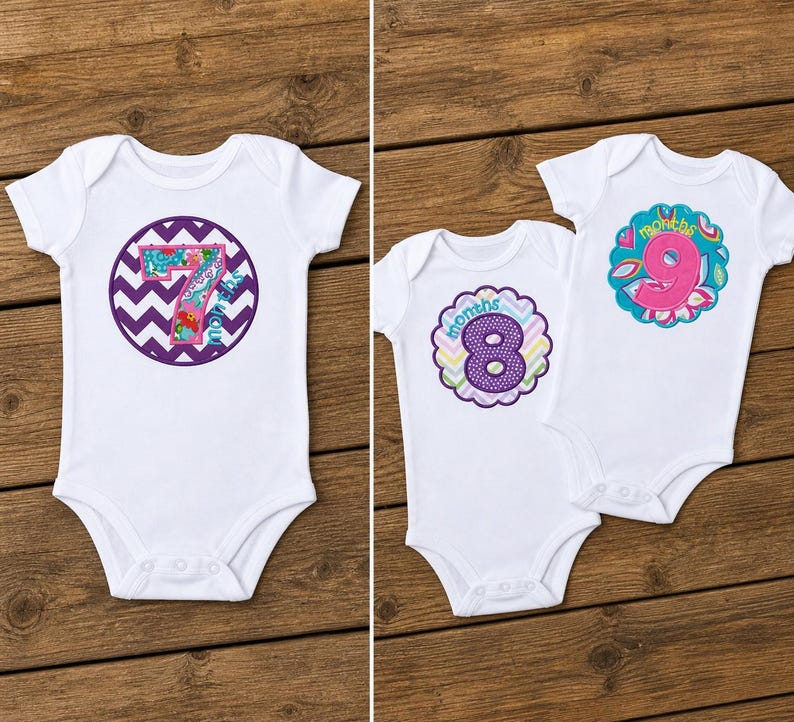 May include: White baby onesies with colorful appliqued numbers 7, 8, and 9, each surrounded by decorative designs. The onesies are displayed on a wooden surface, showcasing the baby clothing.