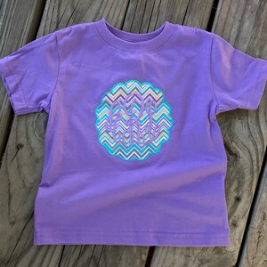 May include: A short-sleeved, lavender t-shirt featuring a decorative, circular emblem with a chevron pattern in shades of blue, gray, yellow, and white. The emblem contains the embroidered initials "A.R.H."