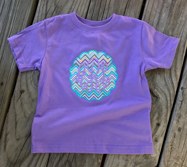 May include: A short-sleeved, lavender t-shirt featuring a decorative, circular emblem with a chevron pattern in shades of blue, gray, yellow, and white. The emblem contains the embroidered initials "A.R.H."