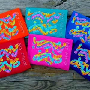 Puede incluir: Seis camisetas coloridas con las palabras "Happy Days" en una fuente tie-dye arcoíris. Las camisetas están todas dobladas y dispuestas en fila sobre una superficie de madera. Cada camiseta tiene las palabras "Play School" impresas en el lateral.