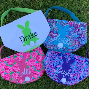May include: Four Easter baskets with bunny designs and personalized names. One basket is white with a green bunny, another has a blue paisley pattern with a blue bunny, a third has a pink floral pattern with a pink bunny, and the last has a purple paisley pattern with a purple bunny.