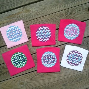 May include: Six colorful t-shirts with embroidered monogram designs. The shirts are pink, white, and light pink. Each shirt features a unique monogram in a scalloped, chevron-patterned circle. The monograms are in shades of blue, green, and purple.