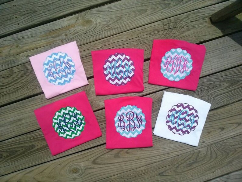 May include: Six colorful t-shirts with embroidered monogram designs. The shirts are pink, white, and light pink. Each shirt features a unique monogram in a scalloped, chevron-patterned circle. The monograms are in shades of blue, green, and purple.