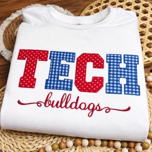 Op de afbeelding: Witte sweatshirt met het woord "TECH" in rode stippen en blauw geruite letters. Het woord "bulldogs" is in rood script eronder geborduurd.