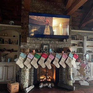 May include: A cozy living room with a brick fireplace adorned with burlap Christmas stockings. A lit fire glows within the hearth. A television is mounted above the fireplace. A small dog rests on the wooden floor.