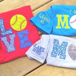 Op de afbeelding: Vier honkbal-thema shirts worden getoond op een houten oppervlak. Een rood shirt heeft het woord "LOVE" met een honkbal en een naam geborduurd op de mouw. Een blauw shirt zegt "T-BALL MOM" met een honkbal.