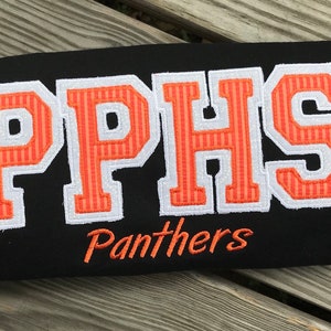 Puede incluir: Artículo rectangular negro con las letras "PPHS" en naranja y blanco, con la palabra "Panthers" bordada debajo. Las letras son de estilo universitario.