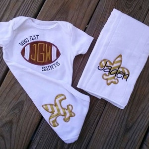 Puede incluir: Conjunto de beb&eacute; blanco con body y manta. El body presenta un dise&ntilde;o de bal&oacute;n de f&uacute;tbol con el texto "WHO DAT SAINTS" y las iniciales "JGW". La manta tiene un dise&ntilde;o de flor de lis y el nombre "Jaden".