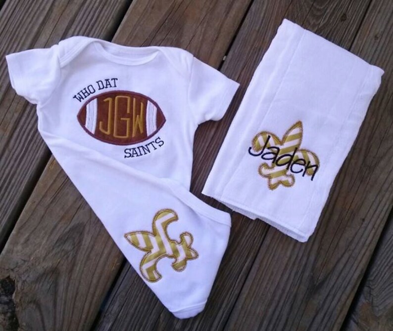Puede incluir: Conjunto de beb&eacute; blanco con body y manta. El body presenta un dise&ntilde;o de bal&oacute;n de f&uacute;tbol con el texto "WHO DAT SAINTS" y las iniciales "JGW". La manta tiene un dise&ntilde;o de flor de lis y el nombre "Jaden".