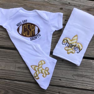 Puede incluir: Un conjunto blanco para beb&eacute; que incluye un body, un babero y un pa&ntilde;o para eructar. El body presenta un bal&oacute;n de f&uacute;tbol bordado con el texto "WHO DAT CSR SAINTS". El babero y el pa&ntilde;o para eructar tienen dise&ntilde;os de flor de lis doradas y el nombre "Charley".