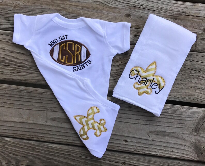 Puede incluir: Un conjunto blanco para beb&eacute; que incluye un body, un babero y un pa&ntilde;o para eructar. El body presenta un bal&oacute;n de f&uacute;tbol bordado con el texto "WHO DAT CSR SAINTS". El babero y el pa&ntilde;o para eructar tienen dise&ntilde;os de flor de lis doradas y el nombre "Charley".