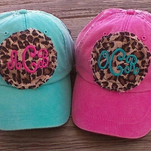 May include: Two baseball caps with a leopard print patch and monogrammed initials. One cap is turquoise and the other is pink.
