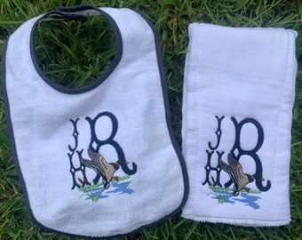 Op de afbeelding: Witte baby-slabbetje en spuugdoek met zwarte geborduurde initialen "J.B." en een eend die over water vliegt.