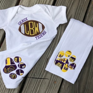 Puede incluir: Conjunto de body y babero para beb&eacute; blancos. El body presenta un dise&ntilde;o de bal&oacute;n de f&uacute;tbol con el texto "GEAUX LBW TIGERS". El babero tiene un dise&ntilde;o de huella de pata con el nombre "Nathan" en morado y dorado.