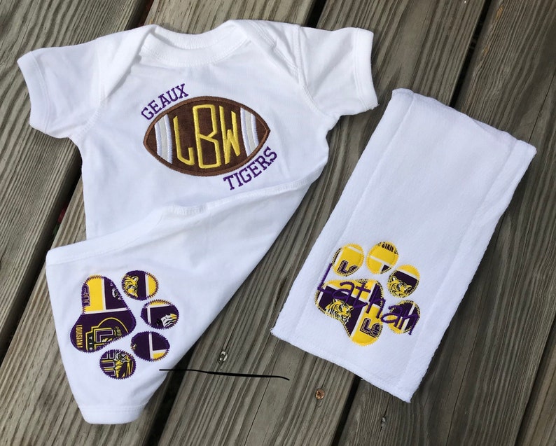 Puede incluir: Conjunto de body y babero para beb&eacute; blancos. El body presenta un dise&ntilde;o de bal&oacute;n de f&uacute;tbol con el texto "GEAUX LBW TIGERS". El babero tiene un dise&ntilde;o de huella de pata con el nombre "Nathan" en morado y dorado.