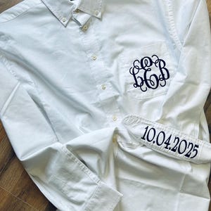 Puede incluir: Camisa blanca de manga larga con botones y un bolsillo en el lado izquierdo. El bolsillo tiene un monograma bordado en hilo negro. El puño de la manga derecha tiene la fecha "10.04.2025" bordada en hilo negro.