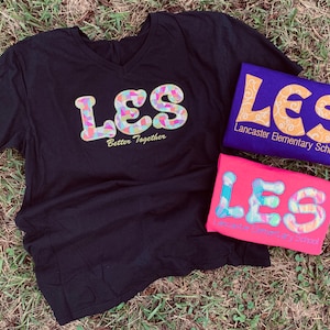 Puede incluir: Tres camisetas con el texto "LES" en letras coloridas y estampadas. La camiseta negra tiene el texto "Better Together" debajo de las letras. La camiseta morada tiene el texto "Lancaster Elementary School" debajo de las letras. La camiseta rosa tiene el texto "Lancaster Elementary School" debajo de las letras.