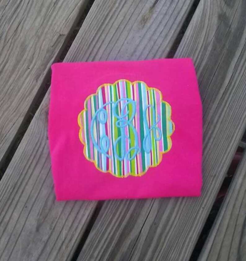 May include: A bright pink t-shirt featuring a scalloped-edge design with colorful vertical stripes and a light blue monogram. The monogram is in a cursive font. The shirt is folded and resting on a wooden surface.