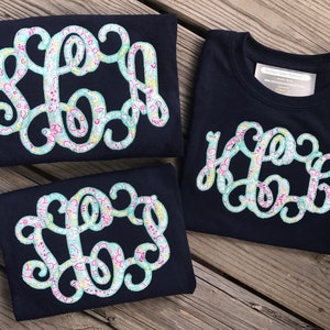 Könnte beinhalten: Drei marineblaue T-Shirts mit einem floralen Monogramm-Design. Das Monogramm ist in einem hellblauen, rosa und gelben floralen Muster.