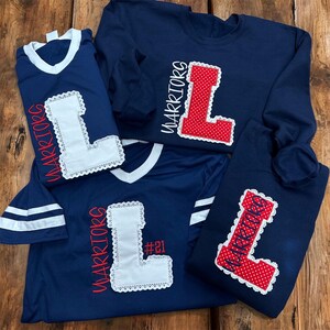 Pode incluir: Uma coleção de camisas e camisolas azul-marinho com aplicações da letra L em branco ou vermelho com bolinhas e acabamento em renda. As peças incluem a palavra WARRIORS bordada em fio vermelho ou branco, e uma camisa exibe o número 21 em vermelho.