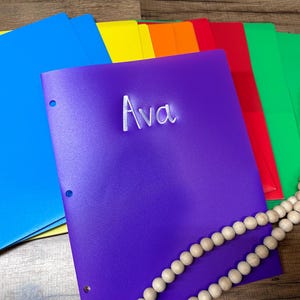 Puede incluir: Una colección de carpetas coloridas en azul, amarillo, naranja, rojo y verde. Una carpeta morada en el centro tiene el nombre "Ava" en escritura blanca. Una guirnalda de cuentas de madera está colocada sobre las carpetas.
