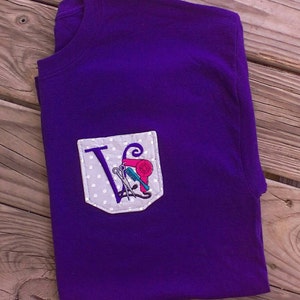 May include: Purple t-shirt with a white pocket featuring a silver letter V and hairdressing tools, including scissors, a comb, and a hairdryer.