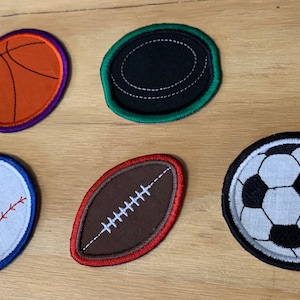 May include: Embroidered patches featuring sports balls: a basketball, hockey puck, baseball, football, and soccer ball. Each patch has a colored border and detailed stitching, perfect for decorating apparel or accessories.
