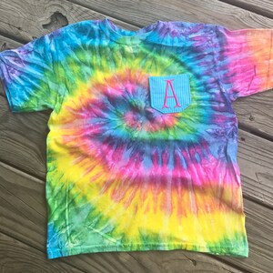 May include: A colorful tie-dye t-shirt with a pocket. The shirt features a spiral pattern in shades of blue, green, yellow, pink, and purple. The pocket is light blue with a pink embroidered letter "A".