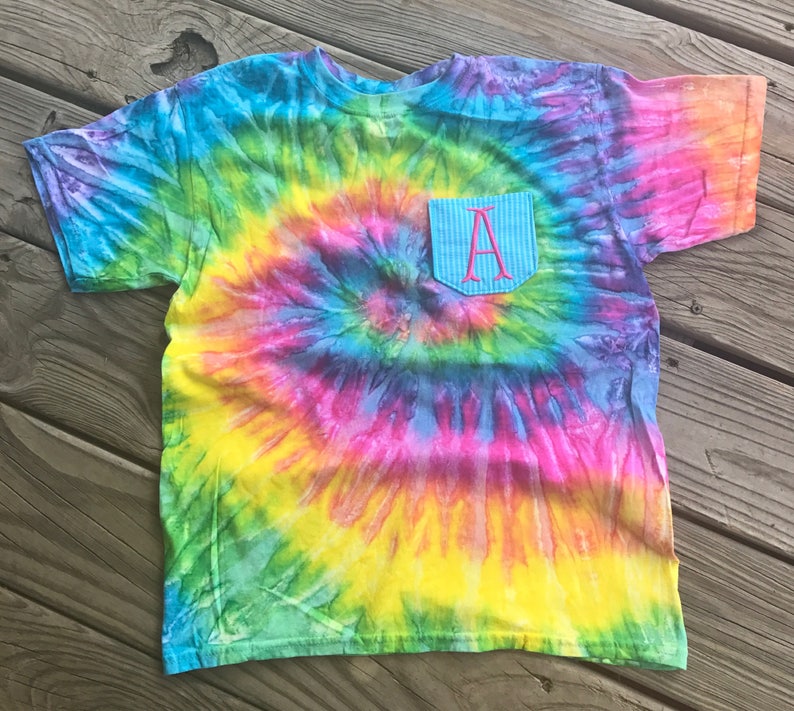 May include: A colorful tie-dye t-shirt with a pocket. The shirt features a spiral pattern in shades of blue, green, yellow, pink, and purple. The pocket is light blue with a pink embroidered letter "A".