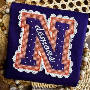May include: A purple shirt featuring an appliquéd letter "N". The letter combines orange gingham and purple polka dots, edged with white lace. The word "demois" is embroidered across the letter.