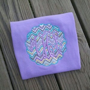 May include: A lavender-colored shirt with a scalloped, patterned applique. The applique features a chevron design in teal, gray, and yellow, with a monogram in a matching lavender color. The shirt is displayed on a wooden surface.
