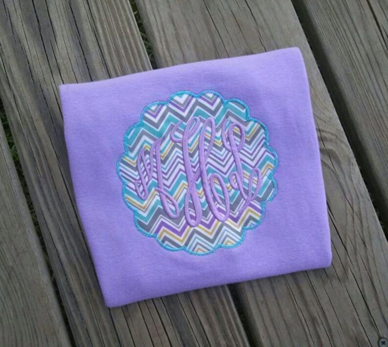 May include: A lavender-colored shirt with a scalloped, patterned applique. The applique features a chevron design in teal, gray, and yellow, with a monogram in a matching lavender color. The shirt is displayed on a wooden surface.