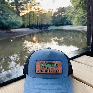 May include: A blue and black trucker hat with a cork patch featuring a green bass fish and the text "HOOKED UP".