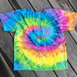 May include: A colorful tie-dye t-shirt with a pocket featuring the letter "K" in pink. The shirt's design includes a spiral pattern of blue, green, yellow, pink, and purple hues. The pocket has a blue and white striped pattern.