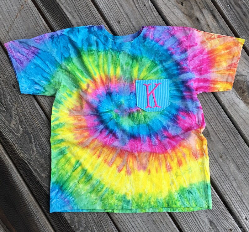 May include: A colorful tie-dye t-shirt with a pocket featuring the letter "K" in pink. The shirt's design includes a spiral pattern of blue, green, yellow, pink, and purple hues. The pocket has a blue and white striped pattern.